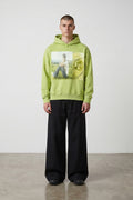 Sudadera con capucha verde lima de Estudio Mora con estampado gráfico de nota musical y persona sentada en un prado.