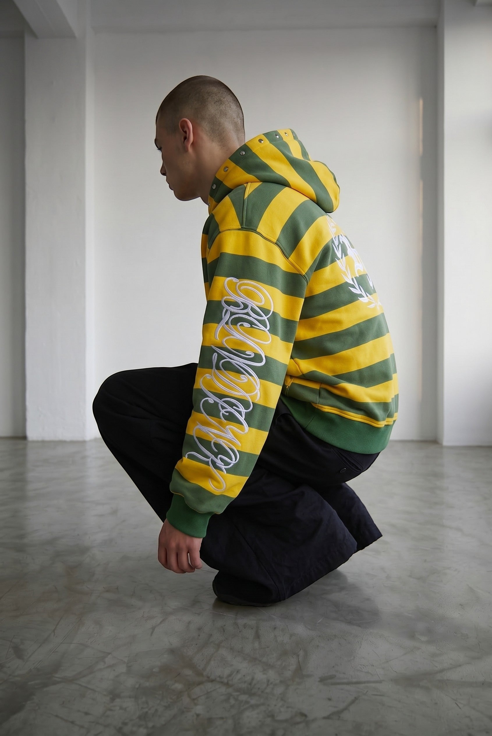 Sudadera con cremallera de Estudio Mora, con rayas verdes y amarillas y bordado blanco en la manga.